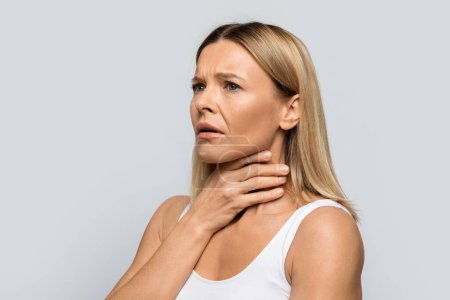 Foto de Sad unhappy adult middle aged blonde woman touching her neck with both hands, lady suffering from sore throat, have pain while swallowing, isolated on grey studio background, looking at copy space - Imagen libre de derechos