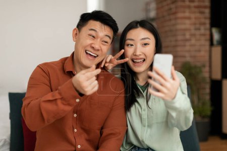Téléchargez les photos : Joyful asian couple enjoying time at home and taking funny selfies together on cellphone, sitting on sofa in living room. Middle aged man and young woman capturing moments on photo - en image libre de droit