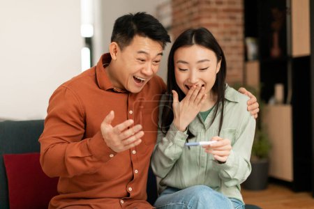 Téléchargez les photos : Happy japanese middle aged husband and his young wife rejoicing positive pregnancy test, sitting in living room interior. Good news, family planning, parenthood and expectation of child - en image libre de droit