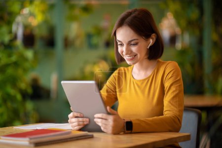 Téléchargez les photos : Cheerful beautiful brunette young lady in casual freelancer working at cafe, sitting at table alone, using wireless earpods, digital pad, have video call with client, copy space. Remote job concept - en image libre de droit