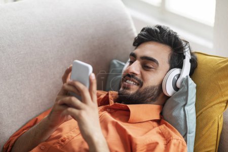 Photo for Cheerful bearded arab guy in casual outfit chilling on couch at home, middle eastern man reclining on sofa, using wireless headphones and cell phone, listening to music, watching content, copy space - Royalty Free Image