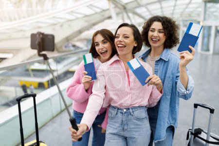 Foto de Tres alegres amigas tomando selfie en el teléfono inteligente en el aeropuerto, mujeres jóvenes felices sosteniendo pasaportes y posando en la cámara, divirtiéndose mientras capturan fotos, disfrutando de viajar juntas - Imagen libre de derechos