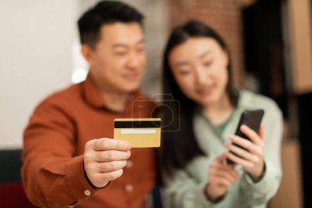 Photo for Asian spouses using smartphone and credit card, shopping online together at home, focus on male hand with debit card. Financial app, banking, sale for shopaholics at home - Royalty Free Image