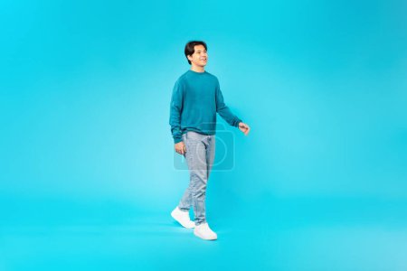 Téléchargez les photos : Happy Japanese Teenager Boy Walking Looking Aside Posing Wearing Casual Clothes Over Blue Studio Background. Teen Age Fashion And Style, Advertisement Banner. Full Length Shot - en image libre de droit