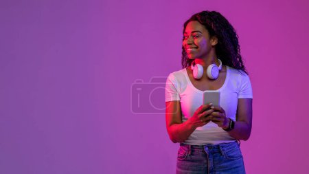 Photo for Smiling Black Female Holding Smartphone And Looking Aside At Copy Space While Standing In neon Light Over Purple Background, Happy African American Lady Enjoying Mobile Offer, Panorama - Royalty Free Image