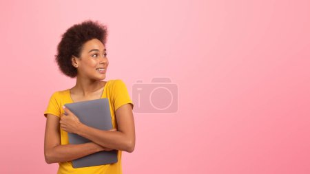 Téléchargez les photos : Joyeux jeune femme afro-américaine bouclée en t-shirt jaune presse ordinateur portable à la poitrine, regarde l'espace vide isolé sur fond rose, studio, panorama. Dispositif pour le travail, étude, chat, annonce et offre - en image libre de droit