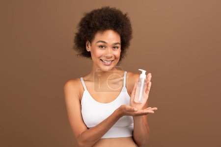 Photo for Cheerful young multiethnic curly female with perfect skin in white top shows jar of lotion, recommends cosmetics, isolated on brown background, studio. Beauty care, moisturizing, anti-aging treatment - Royalty Free Image