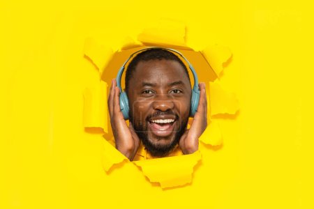 Téléchargez les photos : Homme noir excité touchant écouteurs sans fil et écoutant de la musique en ligne, regardant et souriant à travers un trou dans un fond de papier jaune déchiré. Offre musicale - en image libre de droit