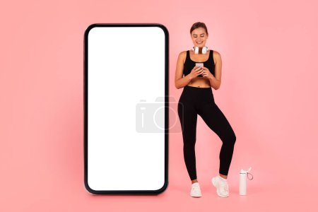 Photo for Happy Sporty Lady With Smartphone In Hands Standing Near Huge Blank Cellphone With White Screen, Smiling Fit Woman In Sportswear Browsing New Fitness App While Standing On Pink Background, Mockup - Royalty Free Image