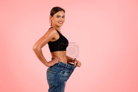 Téléchargez les photos : Beautiful Slim Young Woman Wearing Big Oversize Jeans Posing Isolated Over Pink Studio Background, Happy Millennial Lady Comparing Size, Enjoying Result Of Successful Weightloss, Copy Space - en image libre de droit