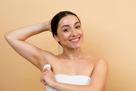 Téléchargez les photos : Personal Hygiene. Smiling Young Indian Woman Applying Stick Deodorant To Underarm Zone, Beautiful Hindu Lady Using Antiperspirant On Armpit To Reduce Sweating, Posing Over Beige Studio Background - en image libre de droit