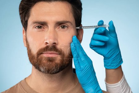 Photo for Closeup shot of handsome bearded man getting filler for eyes zone at beauty salon or aesthetic clinic, blue studio background. Doctor in protective blue gloves injecting filler - Royalty Free Image