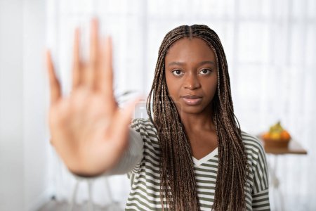 Photo for Serious attractive young black woman with long braids in casual outfit showing palm stop gesture at camera, home interior. Stop domestic abuse, violence concept - Royalty Free Image