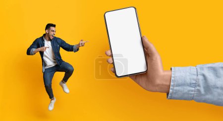 Foto de Cheerful Young Arab Guy Jumping And Pointing At Big Blank Smartphone In Huge Female Hand, Overjoyed Middle Eastern Man Emotionally Reacting To Online Offer Or Mobile Application, Panorama, Mockup - Imagen libre de derechos