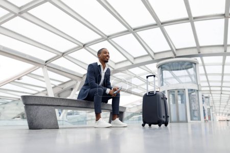 Foto de Air Travels Concept. Smiling African American Male In Suit Sitting On Bench At Airport, Young Black Businessman With Suitcase Waiting For Flight Boarding Near Terminal Gate, Enjoying Business Trip - Imagen libre de derechos