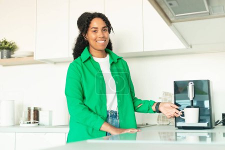 Téléchargez les photos : Cheerful Black Woman Making Espresso Drink Via New Coffee Machine Posing Smiling Looking At Camera Standing In Modern Kitchen At Home On Weekend. Morning Coffee Routine Concept - en image libre de droit