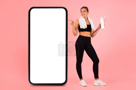 Photo for Cool App. Cheerful Sporty Lady Pointing At Big Blank Smartphone With White Screen, Happy Young Athletic Woman Holding Fitness Bottle And Indicating Copy Space For Advertisement, Mockup - Royalty Free Image