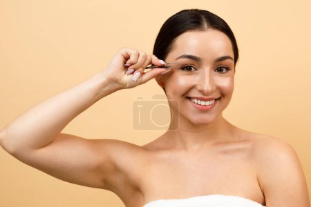 Téléchargez les photos : Portrait Of Beautiful Young Indian Woman Plucking Eyebrows With Tweezers And Smiling At Camera, Happy Hindu Female Making Beauty Self-Care Routine While Standing Over Beige Studio Background - en image libre de droit
