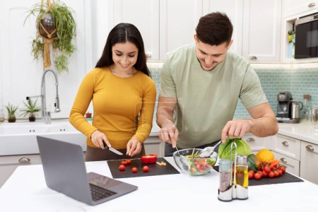 Téléchargez les photos : Positive happy young lovers having fun while cooking at home, cheerful hispanic man and woman preparing delicious healthy meal, watching food vlog on Internet, using laptop. Healthy diet - en image libre de droit