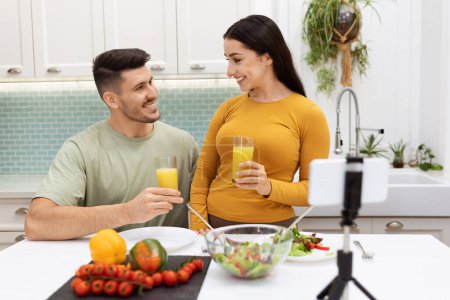 Téléchargez les photos : Cheerful positive beautiful young loving couple food bloggers streaming while have dinner at home, happy brunette man and woman shooting content on smartphone, modern kitchen interior, copy space - en image libre de droit