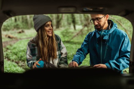 Téléchargez les photos : Happy millennial caucasian guy and woman in jackets take stuff out of car trunk in cold forest, enjoy adventure, inside. Active lifestyle and camping, hikers at vacation, auto travel and relationships - en image libre de droit