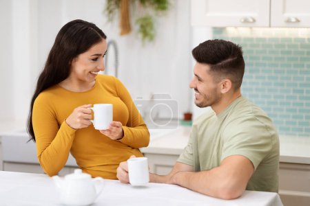 Photo for Young couple in love drinking tea together in kitchen, happy beautiful hispanic man and woman in homewear enjoying morning coffee at home, have conversation, smiling, copy space. Love, relationships - Royalty Free Image
