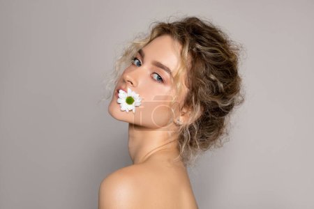 Photo for Attractive young woman holding chamomile in mouth, advertising skin care cleansing products and moisturisers with natural ingredients, posing on grey background - Royalty Free Image