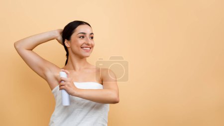 Photo for Portrait Of Smiling Young Indian Woman Using Deodorant Spray For Underarms, Beautiful Hindu Female Wrapped In Towel After Bath Using Antiperspirant Aerosol Over Beige Background, Panorama, Copy Space - Royalty Free Image