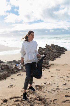 Foto de Día de la Tierra. Joven activista poniendo botellas de plástico en una bolsa de basura, limpiando la zona costera, recortada, tiro vertical. El concepto de conservación ambiental - Imagen libre de derechos