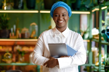 Foto de Portrait of pretty happy young black woman SMM manager wearing smart casual and blue turban posing at cafe while working online, holding digital tablet and notepad, smiling at camera, copy space - Imagen libre de derechos