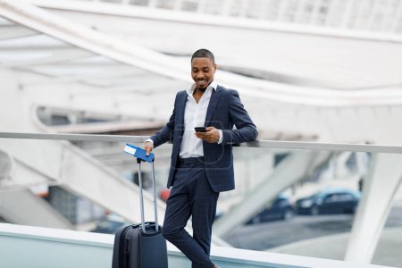 Foto de Hombre de negocios negro sonriente que usa el teléfono inteligente mientras espera el vuelo en el aeropuerto, joven empresario afroamericano con traje que se relaja con teléfono móvil en la terminal, reserva de hotel en línea - Imagen libre de derechos