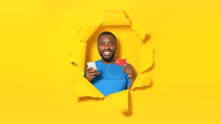 Foto de Hombre afroamericano positivo sosteniendo tarjeta de crédito y utilizando el teléfono celular para el pago electrónico, rompiendo a través de agujero roto en el fondo de papel amarillo. Gestión de la financiación de Internet - Imagen libre de derechos