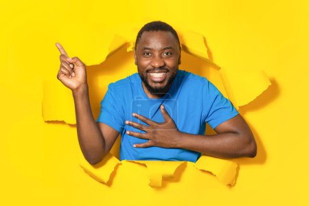 Téléchargez les photos : Homme noir excité pointant de côté à l'espace libre à travers le trou dans le papier jaune déchiré, posant et la publicité offre wow. Concept de publicité commerciale - en image libre de droit