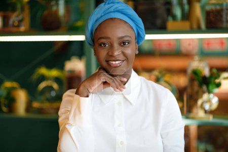 Téléchargez les photos : Portrait rapproché d'une jolie jeune femme afro-américaine joyeuse portant un turban traditionnel posant au café, souriant à la caméra, touchant son visage, copiant l'espace. Millennials concept de style de vie - en image libre de droit