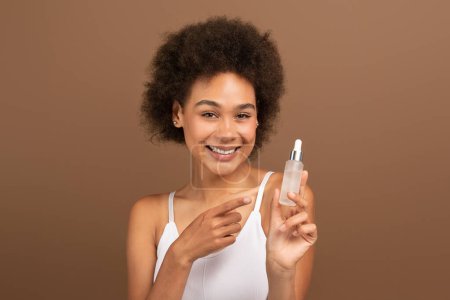 Téléchargez les photos : Smiling young black curly lady with perfect skin point finger at jar of oil, serum, recommend moisturizing isolated on brown background, studio. Ad and offer, cosmetic for skin treatment, beauty care - en image libre de droit
