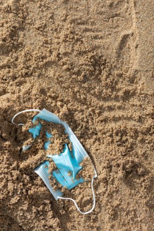 Foto de Mascarilla médica quirúrgica desechada yaciendo en la arena en la playa, primer plano, recortada, tiro vertical. Contaminación plástica, limpieza del océano, COVID-19, residuos médicos, basura, basura en el agua de mar - Imagen libre de derechos