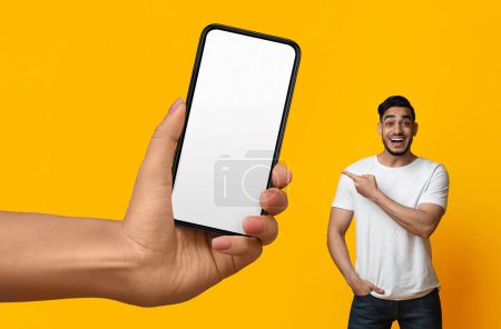Téléchargez les photos : Cheerful arab man pointing at big blank smartphone in giant female hand, happy young middle eastern guy showing copy space for mobile app design or online offer, standing on yellow background, mockup - en image libre de droit