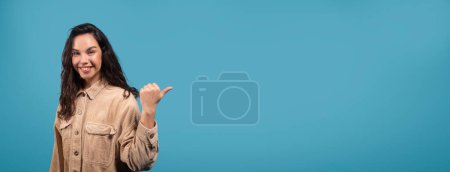 Foto de Feliz bonita milenaria europea estudiante morena apuntando con el dedo al espacio vacío, aislada sobre fondo azul, panorama, estudio. Recomendación y asesoramiento profesional, venta enorme, anuncio y oferta - Imagen libre de derechos