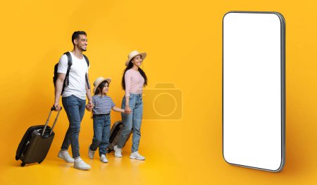Téléchargez les photos : Happy cheerful middle eastern family of three with suitcases going on summer vacation together, parents and little daughter walking with luggage towards phone with mockup on yellow studio background - en image libre de droit