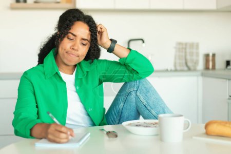 Téléchargez les photos : Black Woman Learning And Having Breakfast Taking Notes Sitting In Modern Kitchen At Home. Pensive Lady Writing Planning Healthy Diet Menu. Dieting And Nutrition Concept - en image libre de droit