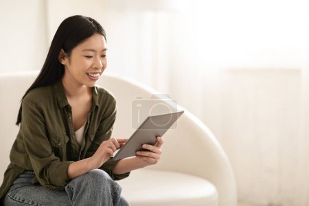 Téléchargez les photos : Joyeux joyeux mignon attrayant coréen dame avec de longs cheveux soyeux assis sur le canapé avec tampon numérique, lecture blog, défilement sur les médias sociaux, regarder le contenu, intérieur de la maison, panorama avec espace de copie - en image libre de droit
