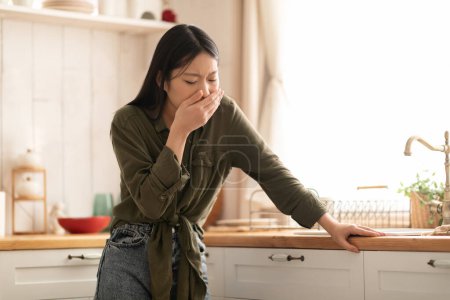 Téléchargez les photos : Jeune femme asiatique dans le sentiment décontracté nausées, couvrir la bouche, debout à côté de la table dans la cuisine, copier l'espace. Toxicose pendant la grossesse, concept d'intoxication alimentaire - en image libre de droit
