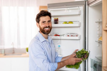 Photo for Glad hungry attractive adult caucasian man with beard takes out fresh salad from refrigerator, enjoy organic meal in light kitchen interior. Lunch at home, proper nutrition, health care, ad and offer - Royalty Free Image