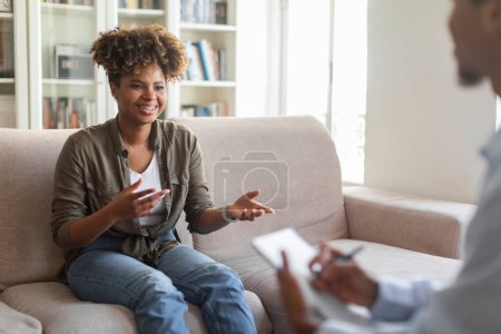 Photo for Happy cheerful young african american woman in casual outfit sitting on couch at cozy counselor office, having conversation with therapist black man, sharing good results of therapy and smiling - Royalty Free Image