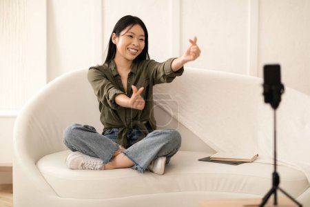 Photo for Cute pretty stylish millennial chinese lady vlogger recording content for followers in social media, sitting on couch and gesturing, looking at cell phone set on tripod and smiling, copy space - Royalty Free Image