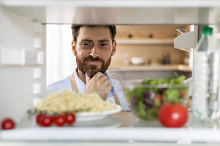 Photo for Glad pensive hungry adult caucasian man with beard looks in refrigerator, chooses pasta or salad in kitchen interior, inside, close up. Diet, proper nutrition, health care, ad and offer, homemade food - Royalty Free Image