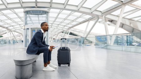 Téléchargez les photos : Transportation Concept. Young Black Businessman With Suitcase Waiting For Flight At Airport, Bored African American Male Sitting On Bench At Terminal Hall, Having Plane Boarding Delay, Copy Space - en image libre de droit