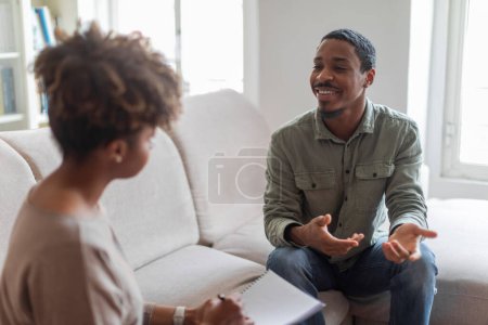 Photo for Cheerful positive handsome young african american guy in casual outfit sitting on couch at cozy counselor office, having conversation with therapist black woman, sharing good results of therapy - Royalty Free Image