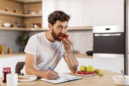 Photo for Busy happy adult caucasian man in white t-shirt eats sandwich with jam, makes notes in notebook at table in kitchen interior. Breakfast and work, study remotely, good morning, weekend home alone - Royalty Free Image