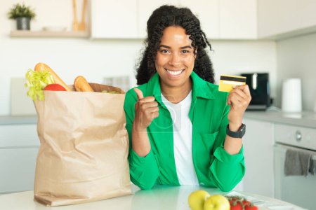 Photo for I Like Online Grocery Shopping. Happy African American Woman Showing Credit Card And Thumbs Up Gesture Posing With Paper Bag Full Of Food Products Standing In Modern Kitchen At Home - Royalty Free Image